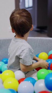 Lovely smiling toddler boy sitting in the dry pool. Baby boy throws the colorful balls to the corridor. Vertical video