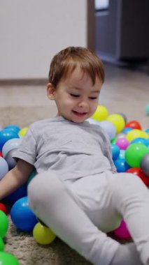 Caucasian toddler lies down on the colorful balls scattered on the floor. Happy active boy has fun time at home. Vertical video