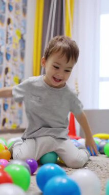 Sevimli erkek bebek yerde oturup el sallıyor. Sevimli çocuk topları odada atıyor. Dikey video