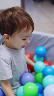 Güzel, koyu saçlı çocuk kuru havuzda oturuyor. Gülümseyen çocuk topları kenarlara fırlatıyor ve kafasına bir tane geçiriyor. Dikey video