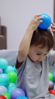 Gri tişörtlü sevimli çocuk kuru havuzda oturuyor. Çocuk mavi bir topu kafasından tutup yere düşürüyor ve el sallıyor. Dikey video