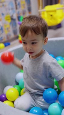 Kuru havuzda oturan sevimli siyah saçlı erkek bebek. Neşeli çocuk topu havuzdan fırlatıyor. Dikey video