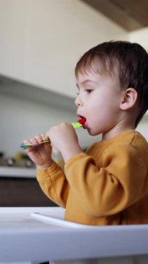 Annem, yüksek sandalyede oturan küçük oğluna hediye paketi veriyor. Tatlı çocuk paketten meyve püresi yiyor. Dikey video