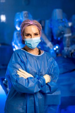 Ameliyathanede kolları kavuşturulmuş kendinden emin bir cerrah. Maskeli ve steril cüppeli kadın cerrah modern ameliyathanede robotik ekipmanlarla çapraz kollarla güven içinde poz veriyor..