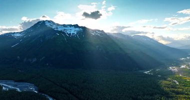 Little cloud covers the sun over the stunning mountain. Rays of sun light the valley covered with pine tree woods. Alaska, USA.