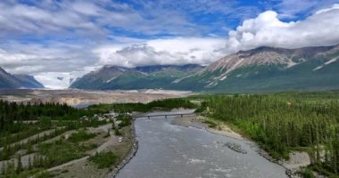 Flying over the turbulent river approaching the bridge above it. Beautiful fluffy cloudscape covers the mountains tops at backdrop. Alaska, USA.