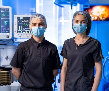 Maskeli iki tıp uzmanı monitörlerin yanında. Siyah üniformalı ve maskeli iki doktor ameliyathanede hasta izleme ekipmanlarının yanında duruyor..