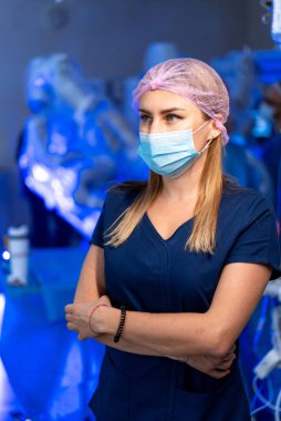 Cerrahi maskeli ve kolları bağlı bir kadın doktor. Ameliyat odasında duran önlüklü, maskeli ve şapkalı kendine güvenen bir kadın doktorun portresi..