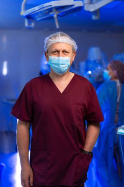 Bordo önlüklü ve maskeli bir erkek doktor ameliyathanede. Burgonya önlüklü ve maskeli bir erkek doktorun portresi. Cerrahi tiyatronun içinde tıbbi malzemelerle..