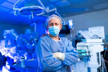 Ameliyat odasında kolları çaprazlanmış önlüklü bir erkek cerrah. Koruyucu cerrahi önlüklü ve maskeli bir erkek doktor bir ameliyathanede tıbbi ekipmanla bekliyor..