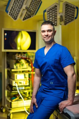 Cerrah ameliyathanede ekipmanla poz veriyor. Ameliyat ışıkları ve tıbbi monitörlerin yanında duran mavi önlüklü doktor..