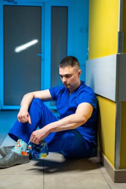 Cerrah hastane koridorunda dinleniyor. Mavi önlüklü doktor ameliyattan sonra duvarın önünde oturuyor..