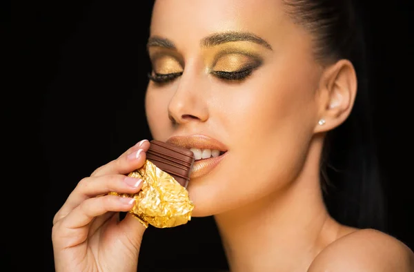 Gorgeous sensual young brunette model eating chocolate wearing golden makeup.