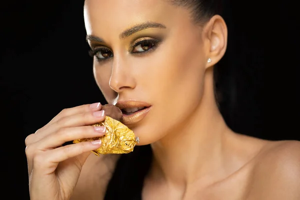 美しい官能的な若いブルネットの女性は黄金の化粧を身に着けてチョコレートを食べる — ストック写真