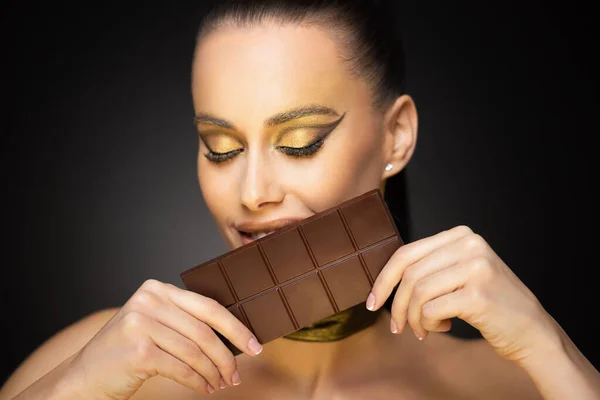 美しい官能的な若いブルネットの女性は黄金の化粧を身に着けてチョコレートを食べる — ストック写真