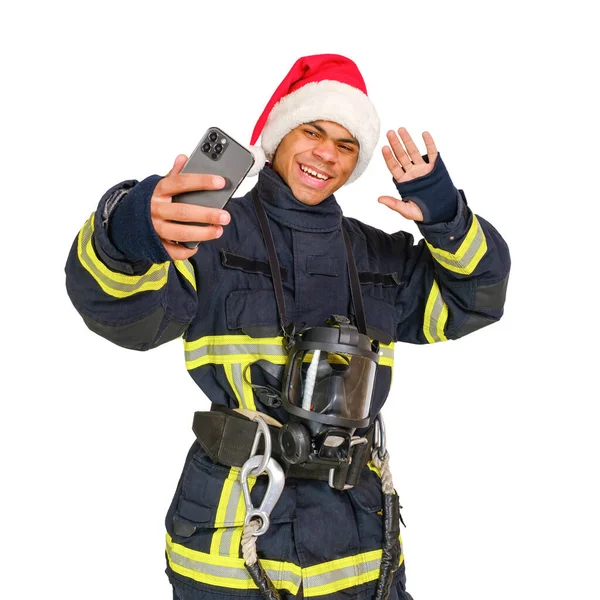 年轻的 面带微笑的非洲裔美国消防员 身穿制服 头戴红色的桑塔爪帽 手里拿着智能手机 把自己隔离在白色背景下 — 图库照片