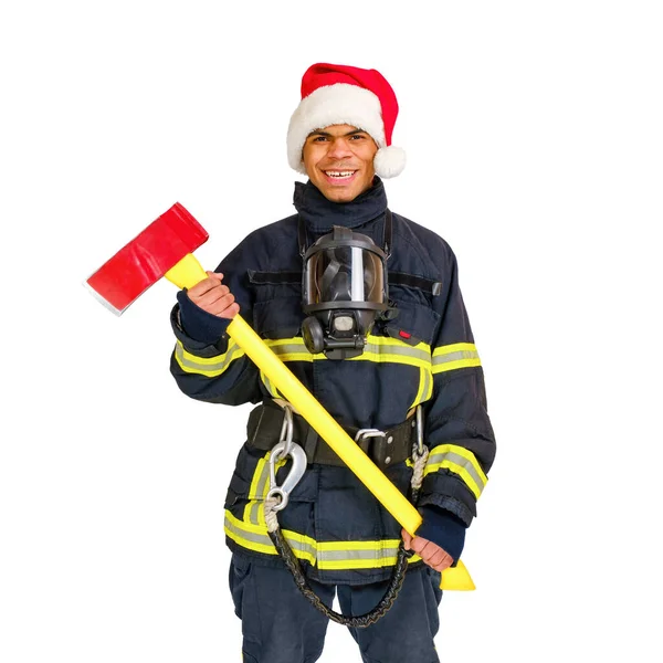 若いアフリカ系アメリカ人の消防士は 耐火制服と赤いサンタの帽子を身に着けており 白い背景に斧を手に持っています孤立 — ストック写真
