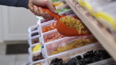 Tek kullanımlık eldivende eli olan kadın bir mağaza çalışanı mağazadaki rafta bir kepçeyle kurutulmuş böğürtlen bırakıyor. Satıcı kadın rafları kuru üzümle dolduruyor, bakkalda çalışıyor..