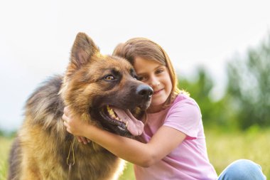 Gülümseyen küçük kız, bir yaz parkında çimlerin üzerinde oturmuş kameraya bakarken çoban köpeğine sarılıyor. Yüksek kalite fotoğraf, yakın görüntü