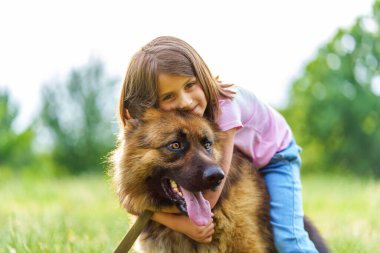 Gülümseyen küçük kız, bir yaz parkında sırt üstü otururken çoban köpeğine sarılıyor. Yüksek kalite fotoğraf, yakın görüntü