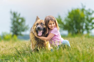 Gülümseyen küçük kız, bir yaz parkında çimlerin üzerinde oturmuş kameraya bakarken çoban köpeğine sarılıyor. Yüksek kalite fotoğraf