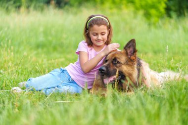 Gülümseyen küçük bir kız çoban köpeğini okşuyor yazın parkta çimlerin üzerinde oturuyor. Yüksek kaliteli fotoğraf, bulanık arkaplan