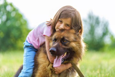 Küçük kız, bir yaz parkında çimlerin üzerinde sırtüstü otururken çoban köpeğine sarılıyor. Yüksek kalite fotoğraf, yakın görüntü