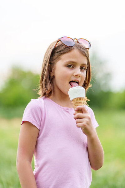 Cute little girl eating an ice cream cone holding it with both hands in summer in the park. High quality photo, blurred background, vertical orientation