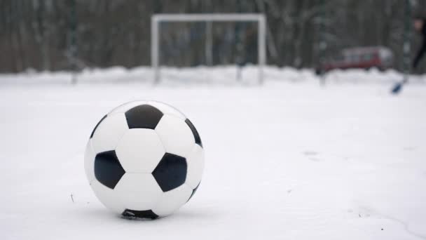 足球位于被雪覆盖的迷你足球场中间的球门前面 而在球门后面 一个身份不明的人拿着一把雪铲清扫球场 — 图库视频影像