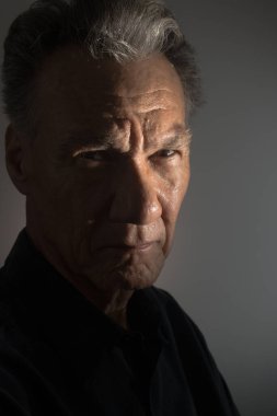 Siyah gömlek giyen, loş ışıkta yaşlı bir adamın kasvetli portresi.