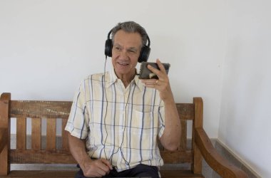 Olgun adam bankta oturmuş akıllı telefonunda kulaklık takmış bir program izliyor.