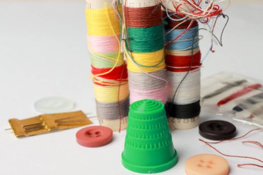 Beyaz zemin üzerinde farklı renk iplik ve iğne makaraları, yüksük ve düğmeler ile seyahat seti