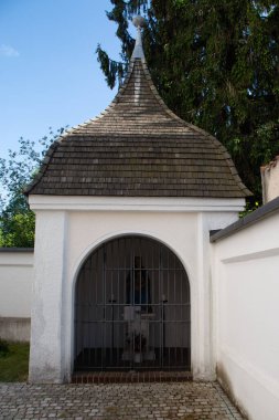 EBERSBERG, GERMANY - 8 Mayıs 2018: Ebersberg 'deki Sankt Sebastian kilisesinin yanında kemerli girişi ve kiremitli çatısı olan küçük beyaz şapel veya türbe, ağaçlar ve açık gökyüzü çerçeveli. Yüksek kalite fotoğraf