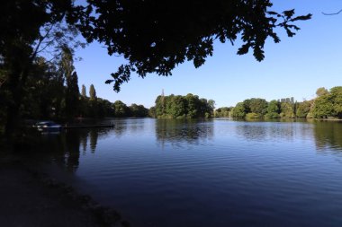 MUNICH, Almanya - 24 Haziran 2025: Eisbach Nehri 'nin İngiliz Bahçesi' nden ağaçlar ve sakin su üzerindeki yansımaları ile huzurlu bir sabah manzarası. Yüksek kalite fotoğraf