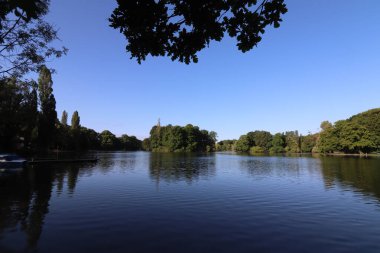MUNICH, Almanya - 24 Haziran 2025: Eisbach Nehri 'nin İngiliz Bahçesi' nden ağaçlar ve sakin su üzerindeki yansımaları ile huzurlu bir sabah manzarası. Yüksek kalite fotoğraf