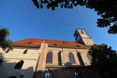 ChatGPT şunları söyledi: WASSERBURG AM INN, GERMANY - 20 Haziran 2025: Wasserburg am Inn 'deki ağaç dalları tarafından çerçevelenmiş öğleden sonra güneş ışığı ve kırmızı çatısı ile St. Jakob kilisesinin alçak açılı görüntüsü. Yüksek