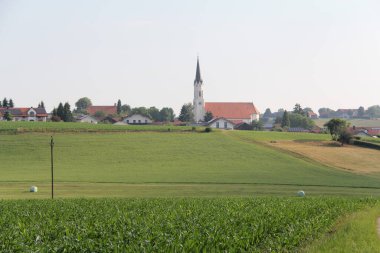 KIRCHDORF, GERMANY - 19 Haziran 2025: Bavyera kırsal kesiminde kırsal alanların üzerinde yükselen ince çan kulesiyle St. Martin Katolik kilisesine uzak bakış. Yüksek kalite fotoğraf
