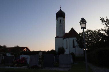 SOYEN, GERMANY - 11 Haziran 2025: Soyen 'deki St. Michael Katolik Kilisesi' nin alacakaranlık manzarası aydınlık cephe ve açık bir akşam gökyüzü altında çevreleyen binalar. Yüksek kalite fotoğraf