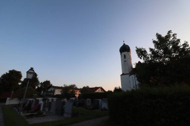 SOYEN, GERMANY - 11 HAZİRAN 2025: Akşam manzaralı St. Michael kilise kulesi ve Soyen köyündeki mezarlık ağaçlardan siluetler ve açık alacakaranlık gökyüzü. Yüksek kalite fotoğraf