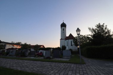 SoYEN, GERMANY - 11 Haziran 2025: Aziz Michael Kilisesi, güneş batarken Bavyera 'nın Soyen köyünde soğan kubbesi ve mezarlıkla, fener ve ağaçlarla döşenmiş asfalt yoldan görüldü. Yüksek kalite fotoğraf