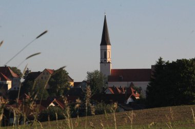 HAAG OBERBAYERN, ALMANY - 31 Mayıs 2025: Oberbayern 'deki Haag kilisesinin uzun uçlu çan kulesi güneş doğarken tarihi köyün çatılarından yükseliyor. Yüksek kalite fotoğraf