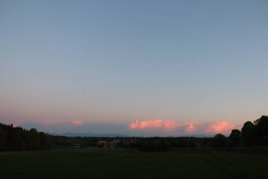 HAAG OBERBAYERN, ALMANY - 29 Nisan 2025: Oberbayern 'deki Haag yakınlarındaki bir ormanlık alanda gün batımında dramatik pembe bulutlar altında Alpler' in uzak görüntüsü. Yüksek kalite fotoğraf