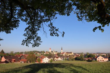 HAAG in OBERBAYERN, GERMANY - 29 Nisan 2025: Oberbayern 'deki Haag' ın ağaç dalları tarafından çerçevelenmiş sabah görüşü, açık mavi gökyüzü altındaki kilise kulelerini ve çatıları gösteriyor. Yüksek kalite fotoğraf