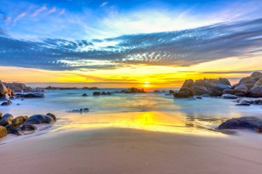 Şafak vakti kumsalda parlak renkli bulutlar, güzel şekilli bir kayanın altında güzel bir manzara yaratıyor.