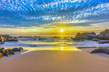 Şafak vakti kumsalda parlak renkli bulutlar, güzel şekilli bir kayanın altında güzel bir manzara yaratıyor.