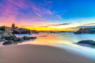 Şafak vakti kumsalda parlak renkli bulutlar, güzel şekilli bir kayanın altında güzel bir manzara yaratıyor.
