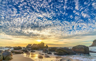 Şafak vakti kumsalda parlak renkli bulutlar, güzel şekilli bir kayanın altında güzel bir manzara yaratıyor.