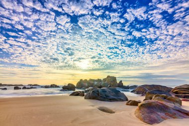 Şafak vakti kumsalda parlak renkli bulutlar, güzel şekilli bir kayanın altında güzel bir manzara yaratıyor.