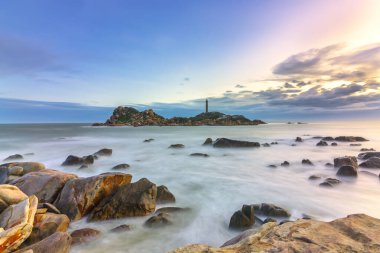 Şafak vakti kumsalda parlak renkli bulutlar, güzel şekilli bir kayanın altında güzel bir manzara yaratıyor.