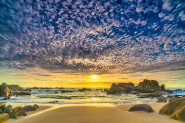 Şafak vakti kumsalda parlak renkli bulutlar, güzel şekilli bir kayanın altında güzel bir manzara yaratıyor.